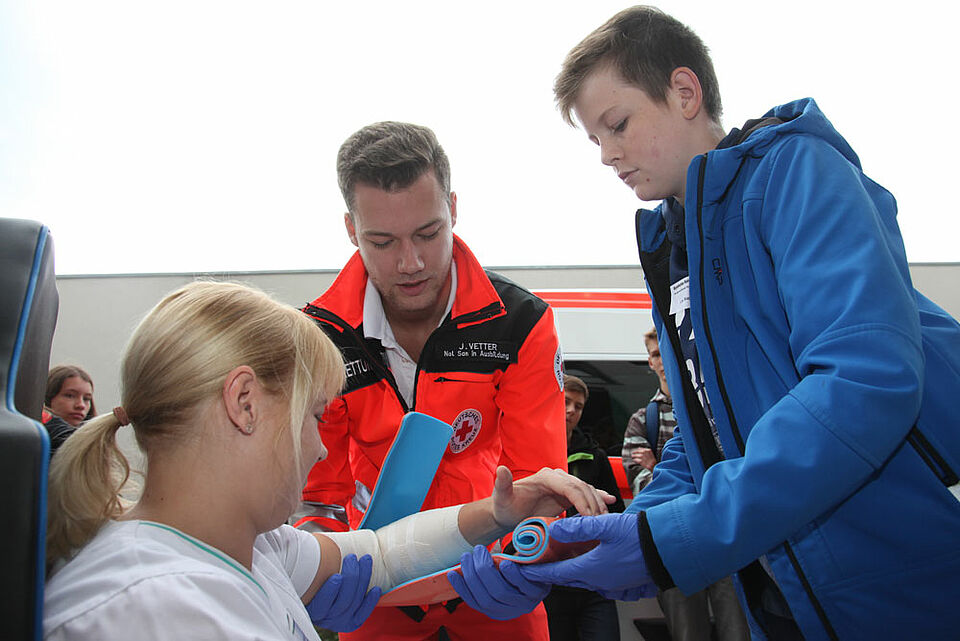 Julian Vetter (Mitte), Auszubildender zum Notfallsanitäter beim BRK, zeigt mithilfe von Jenny Ploner (links), Praxisanleiterin an den Haßberg-Kliniken, die Ruhigstellung einer Armfraktur. (Foto: Michael Will / BRK)